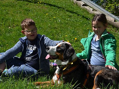 ein Mädchen und ein Junge sitzen auf der Wiese und streicheln einen größeren Hund