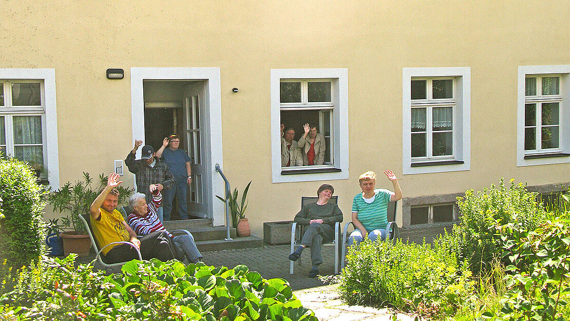 Mehrere Menschen sitzen und stehen vor einem Hauseingang. Und 2 Menschen schauen aus dem Fenster. Einige von Ihnen winken.
